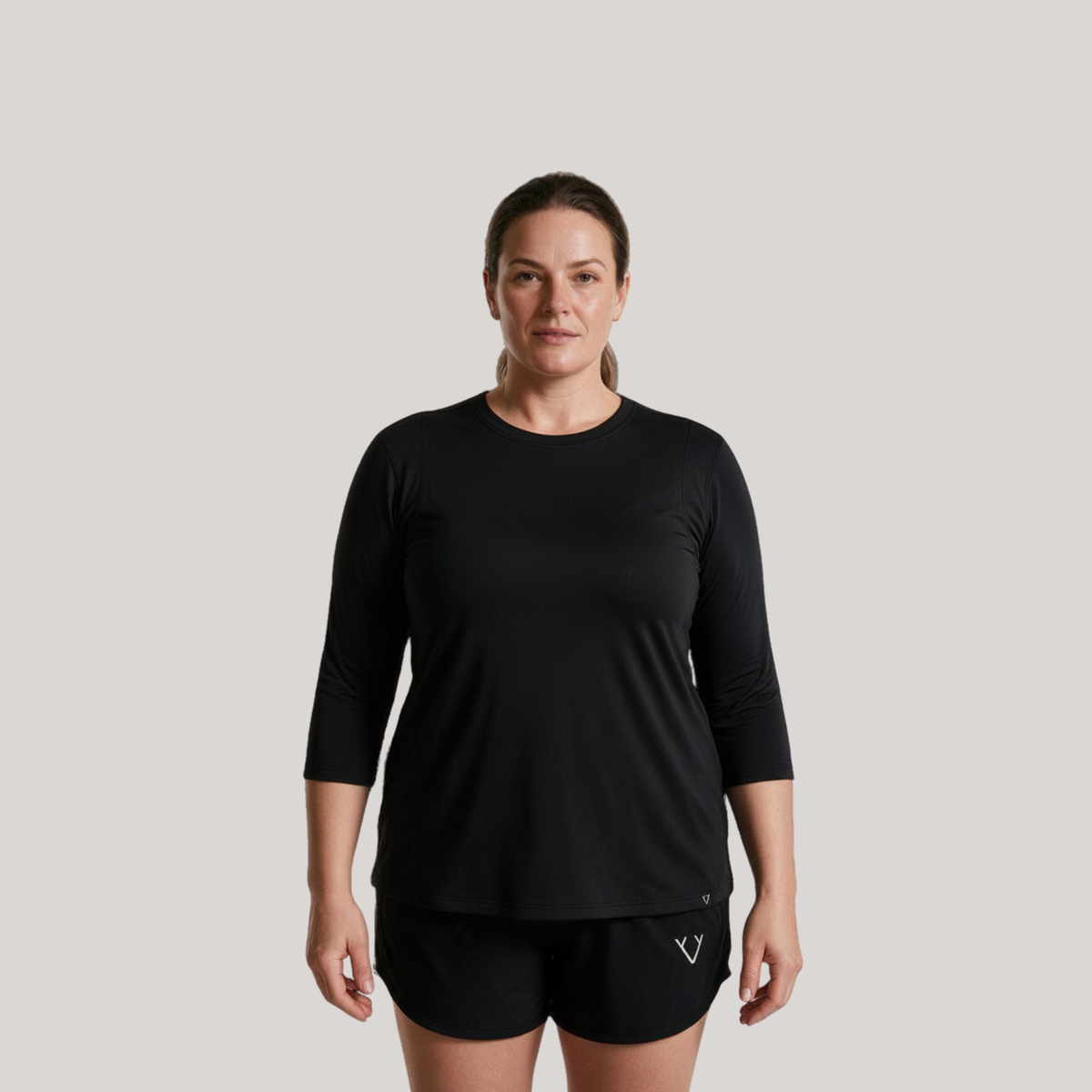 A woman modeling a black three-quarter sleeve athletic top and matching black shorts against a solid light gray background.