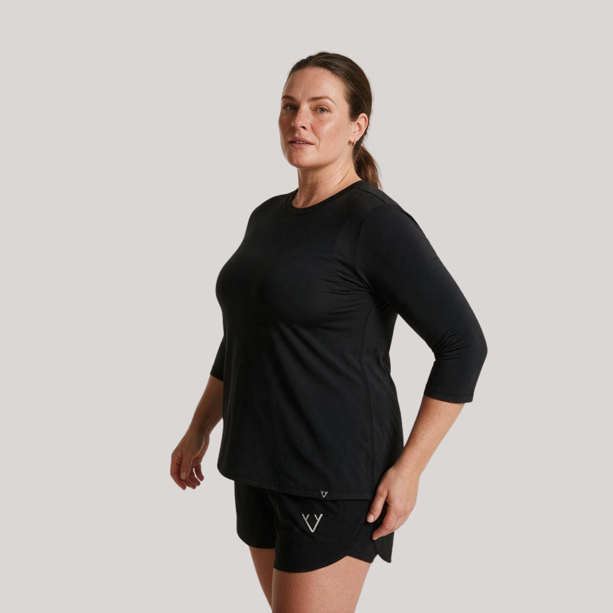 A woman modeling a black three-quarter sleeve athletic top and matching black shorts against a solid light gray background.